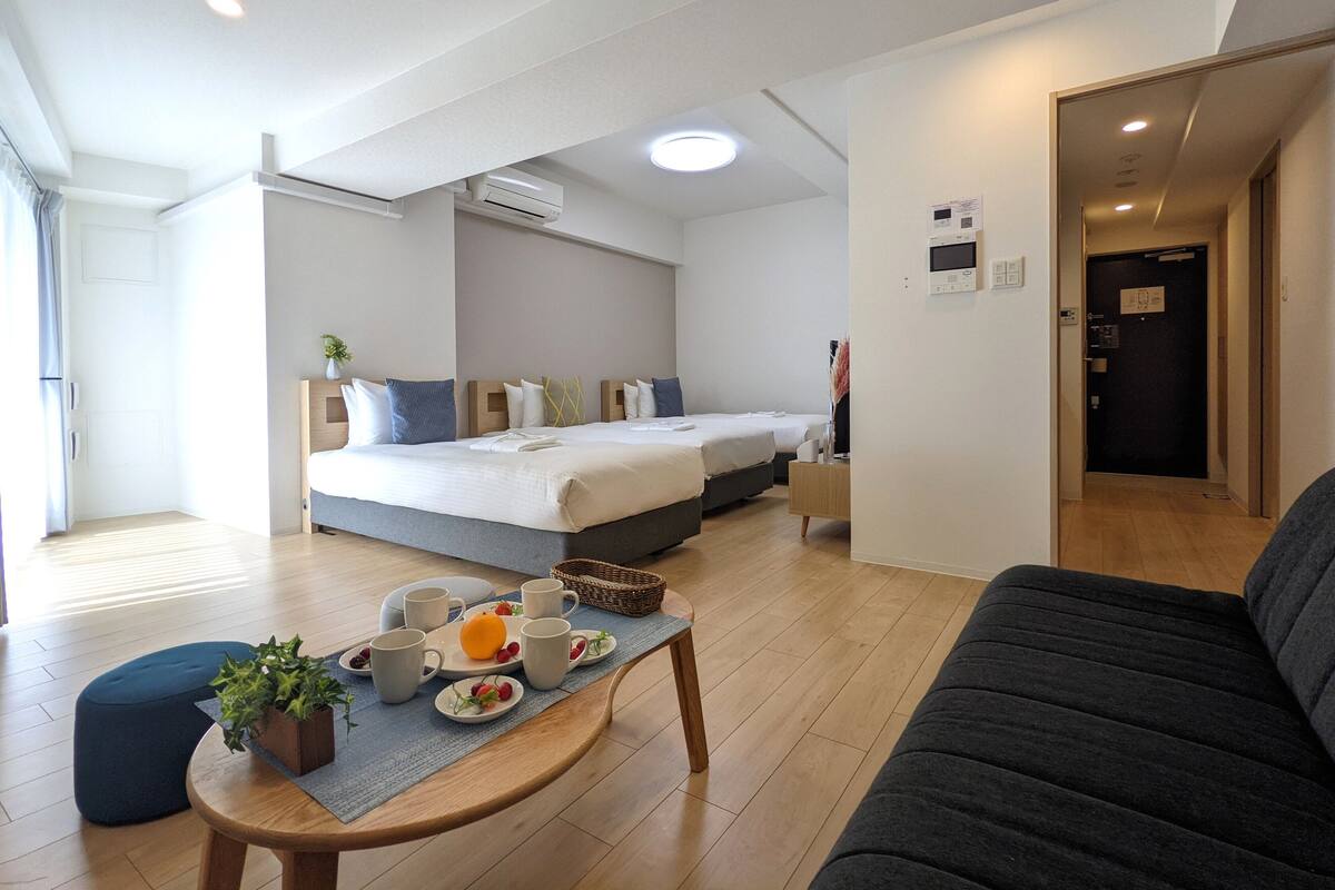 A modern hotel room with a large bed, a sofa, a dining table with fruit and cups, and a small side table.