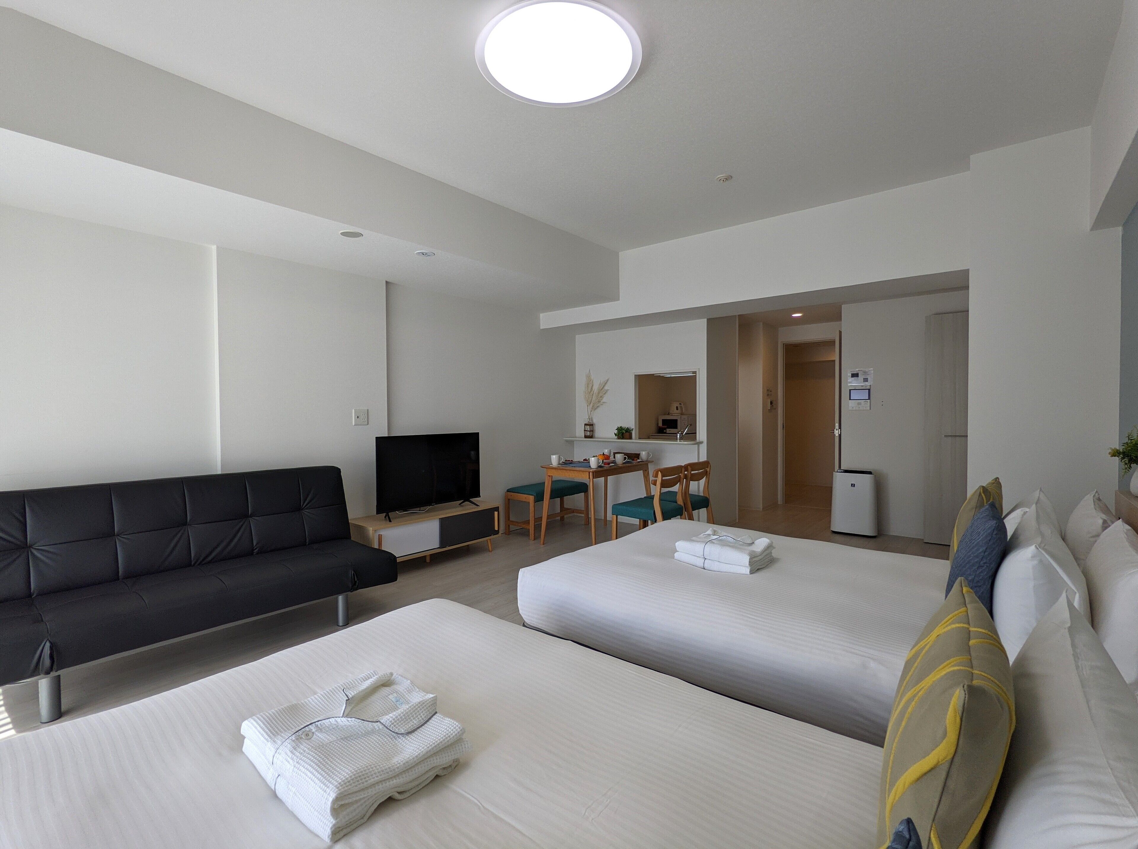 Une chambre d’hôtel moderne avec un canapé noir, deux lits, une télévision, un bureau avec une chaise et une kitchenette.