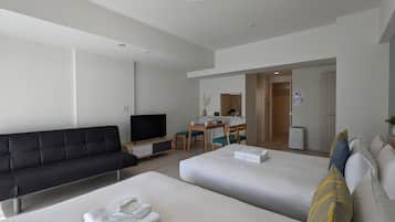 Une chambre d’hôtel moderne avec un canapé noir, deux lits, une télévision, un bureau avec une chaise et une kitchenette.