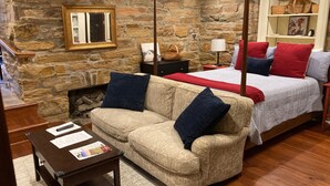 A cozy room with a stone wall, a bed with red and blue bedding, a beige sofa with blue cushions, and a wooden table with papers.