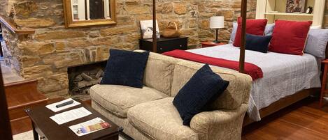 Una habitación acogedora con pared de piedra, una cama con ropa de cama roja y azul, un sofá beige con cojines azules y una mesa de madera con papeles.