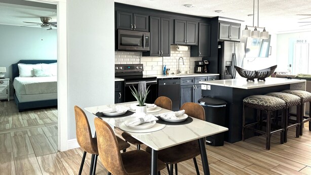 A modern kitchen with dark cabinets, a central island, and a dining area with wooden chairs. - Modern Peaceful Pool Home, 25 Mins To Theme Parks! (St. Cloud)