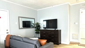 A living room with a grey sofa, a television on a dark wooden cabinet, and a large mirror on the wall. - Modern Peaceful Pool Home, 25 Mins To Theme Parks! (St. Cloud)