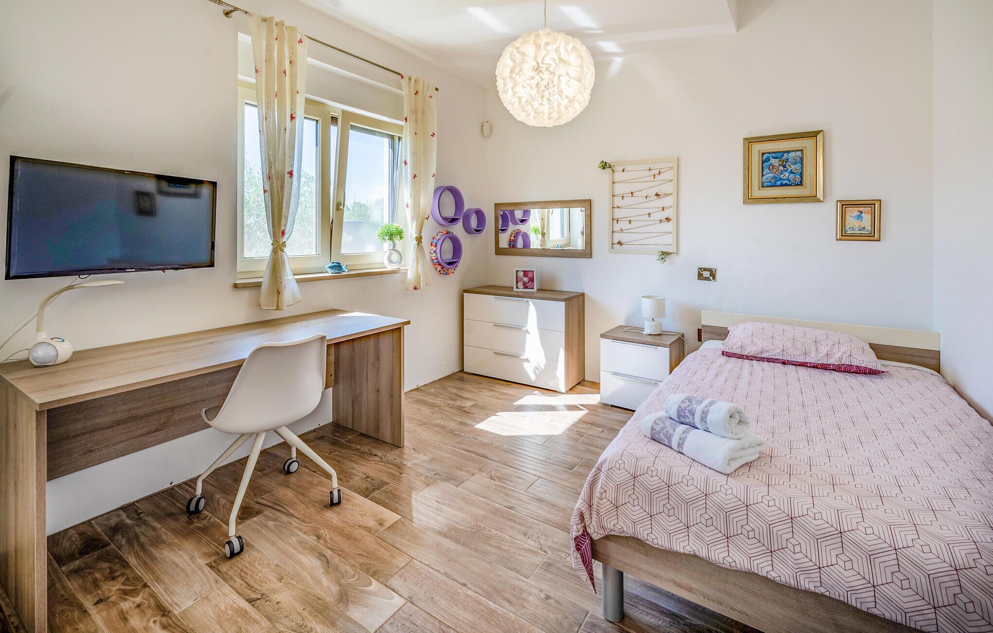 A bedroom with a bed, a desk, a chair, a TV, and a window with curtains.