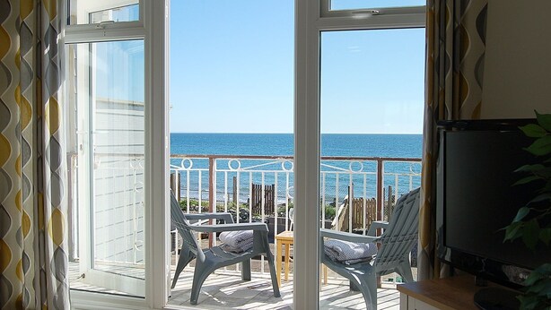 A room with a large sliding glass door leading to a balcony with a view of the sea. There are two chairs on the balcony, and a television is visible on the right side. - Sunny Side Up Beach House in Tywyn (Tywyn)