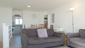 A modern living room with a grey sofa, a white dining table, and a kitchen in the background. - Sunny Side Up Beach House in Tywyn (Tywyn)