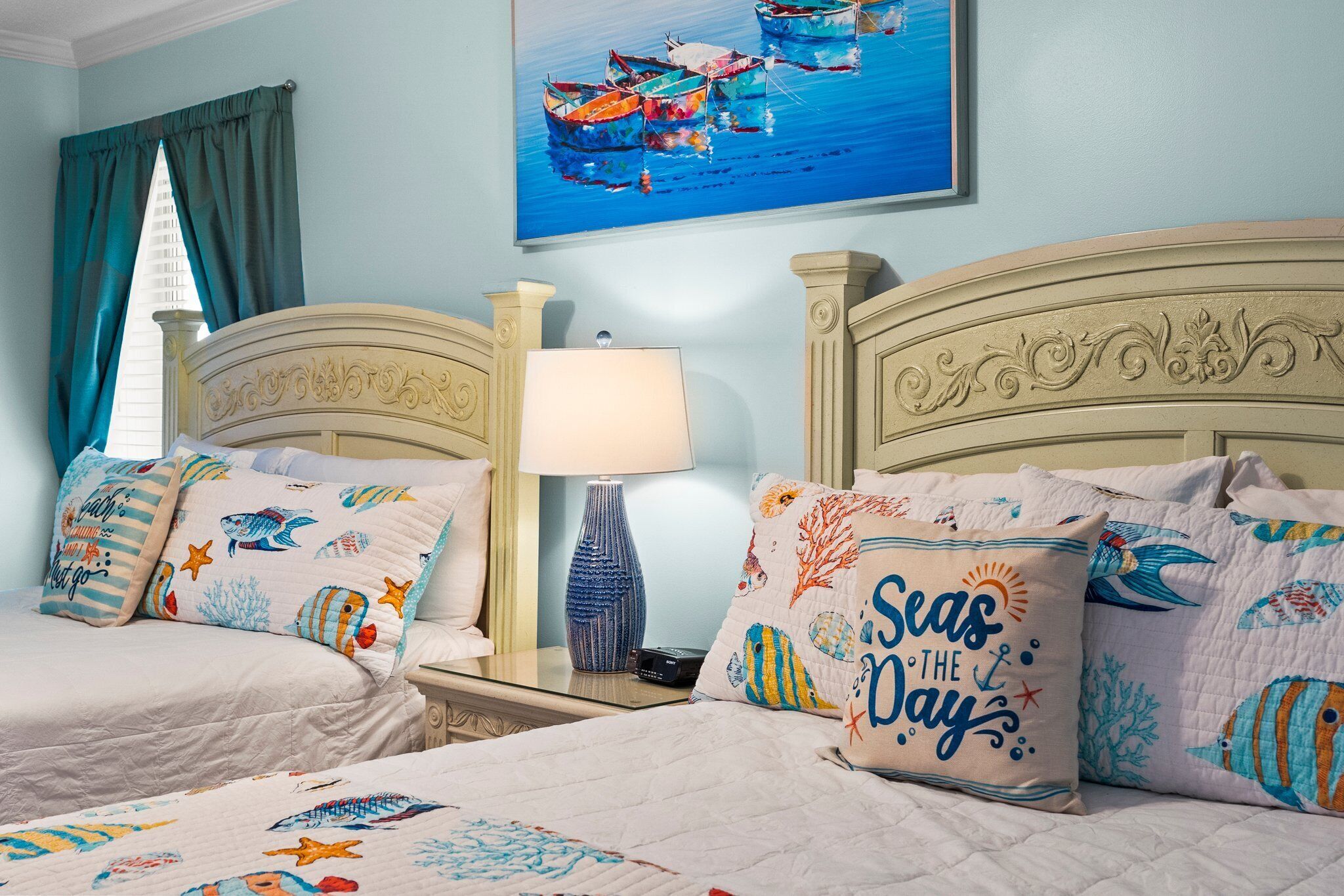 Ein Schlafzimmer mit zwei Betten, jeweils mit einem Bezug und passenden Kissen im maritimen Stil. Ein Nachttisch mit Lampe und ein Bild von Booten an der Wand.