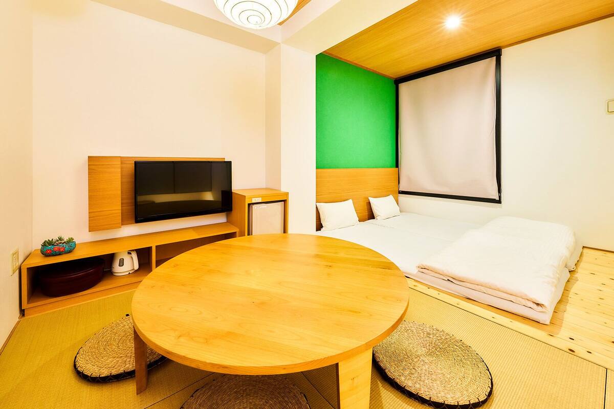 Kompakti hotellihuone, jossa on puinen ruokapöytä, sänky, jonka päätyseinä on maalattu vihreällä, televisio ja pieni keittonurkkaus.