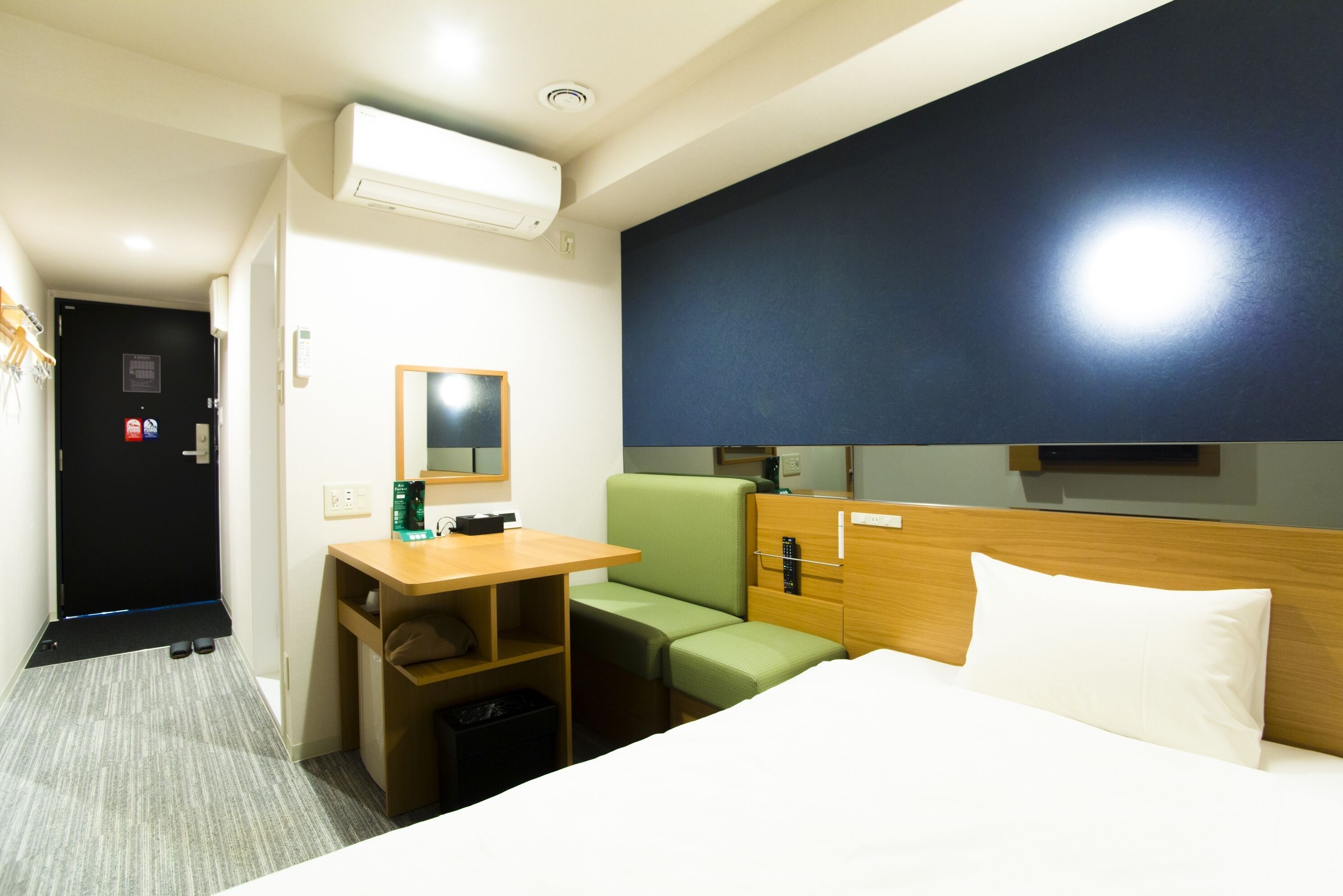 A hotel room with a bed, a desk, a green chair, a wooden headboard, a mirror, and an air conditioning unit.