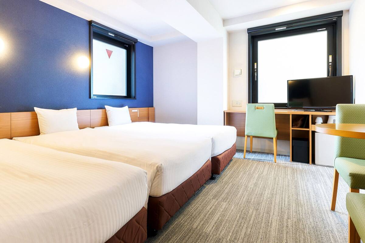 Habitación de hotel con dos camas, un escritorio con televisor y una ventana con persianas.