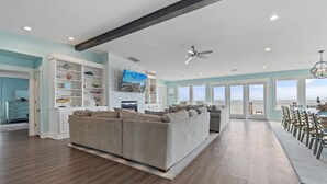 A spacious living room with a large sectional sofa, a dining area with chairs, and a view of the ocean through large windows. - "Happily Ever After" Luxury Oceanfront home w/ Private Pool, 10 Bedrooms, 10.5 Baths (Holden Beach)