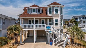 Una casa sulla spiaggia su due piani, con un balcone, un portico coperto e una scalinata che conduce al piano superiore.