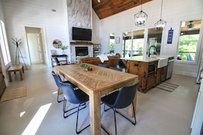 Een ruime keuken met een houten tafel en stoelen, een open haard en een groot raam met uitzicht op groen.