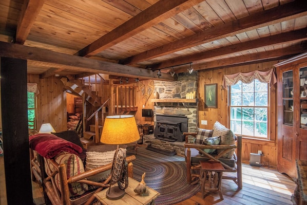 L’intérieur chaleureux d’un chalet avec un plancher en bois, une cheminée, un fauteuil à bascule, une lampe et un escalier menant à un étage supérieur.