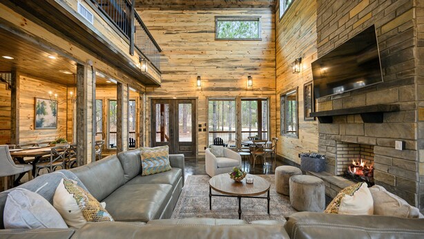 A spacious living room with leather sofas, a stone fireplace, and large windows offering a view of the outdoors. - Sunlight Creek in Broken Bow, Sleeps 20! Creekside & Playset (Broken Bow)