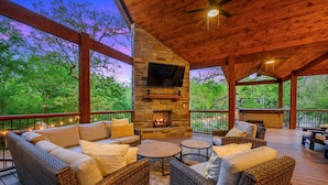 A covered outdoor seating area with wicker furniture, a stone fireplace, and a TV mounted on the wall. - Sunlight Creek in Broken Bow, Sleeps 20! Creekside & Playset (Broken Bow)