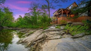 A rustic cabin with a wooden exterior, situated by a rocky riverbank with trees. - Sunlight Creek in Broken Bow, Sleeps 20! Creekside & Playset (Broken Bow)