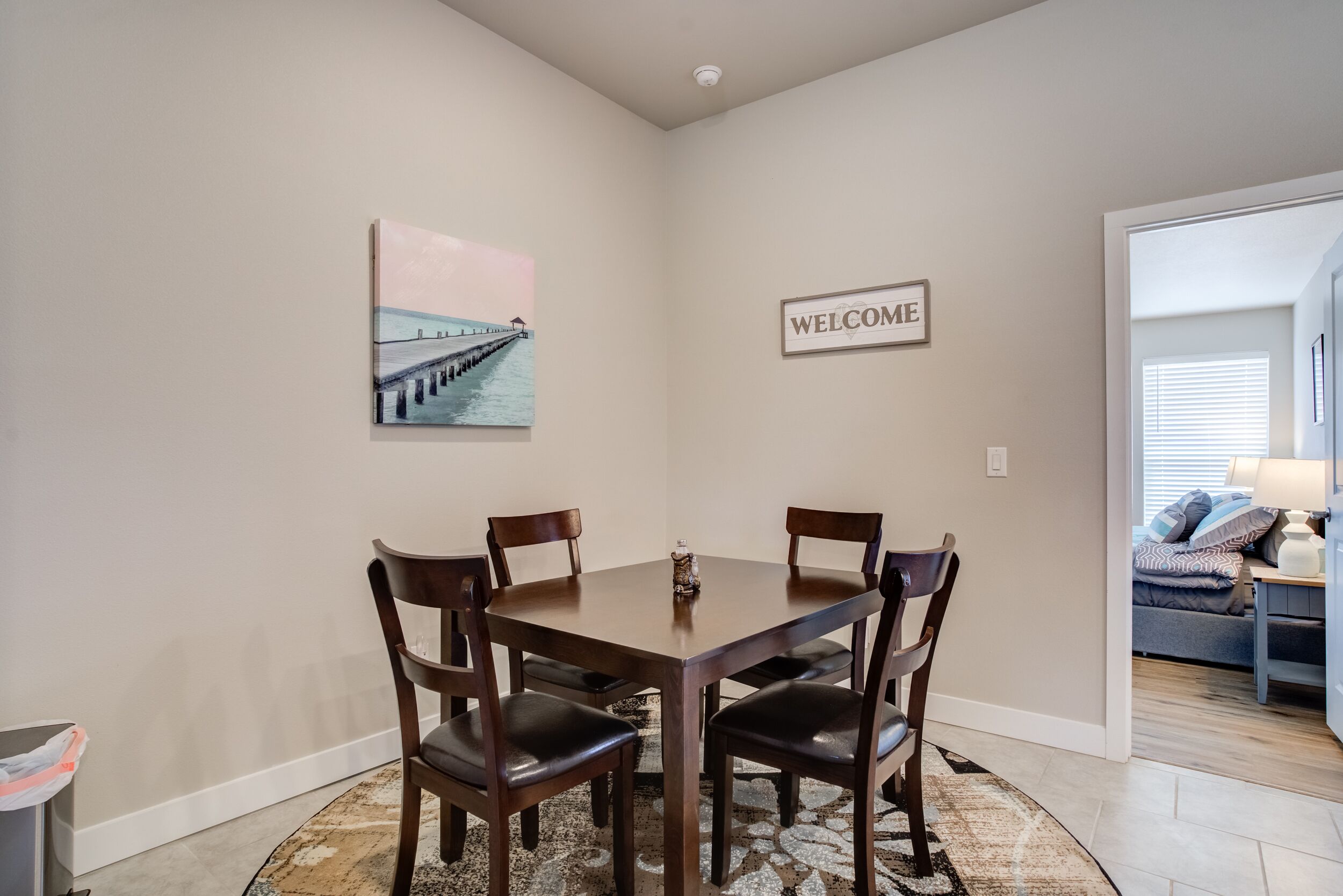 Un coin repas avec une table ronde, quatre chaises, un panneau d’accueil et un tableau représentant une jetée.