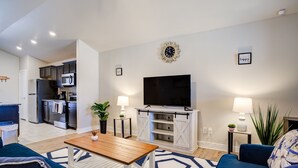 A modern living room with a TV, sofa, coffee table, and kitchen in the background. - Milano Guesthouse (Lubbock)