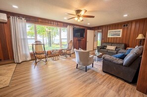 Un salon spacieux doté d’un plancher en bois, d’un ventilateur de plafond, d’une grande fenêtre avec des rideaux, d’un canapé, d’une table basse et d’un téléviseur fixé au mur.