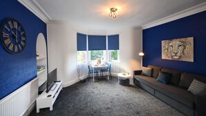 Une pièce avec un mur d’accent bleu, une table ronde avec des chaises, un canapé gris, un téléviseur posé sur un meuble et un tableau accroché au mur.