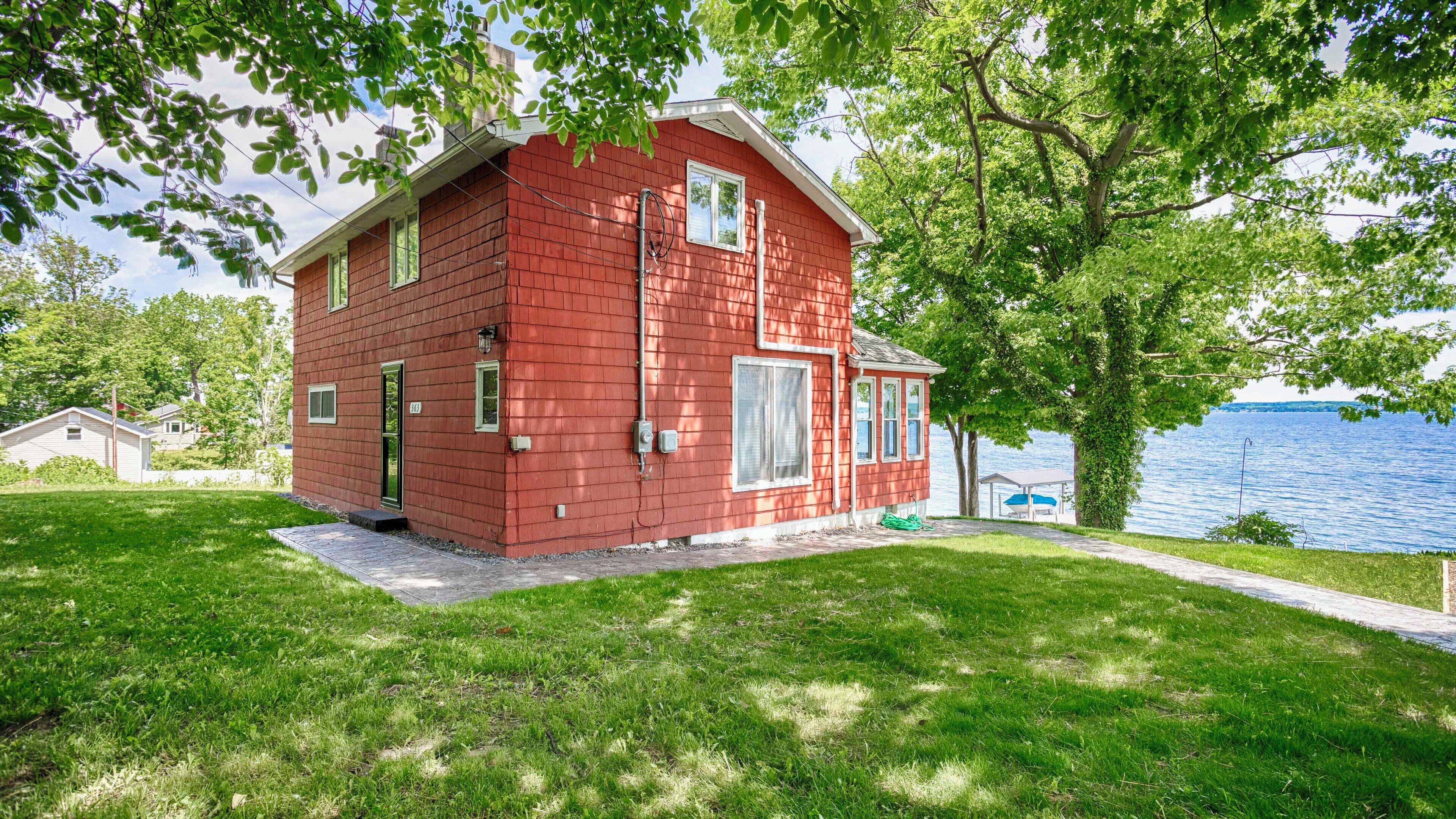 A red two-story house with a front porch, a small deck, and a view of a lake.