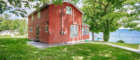 A red two-story house with a front porch, a small deck, and a view of a lake.