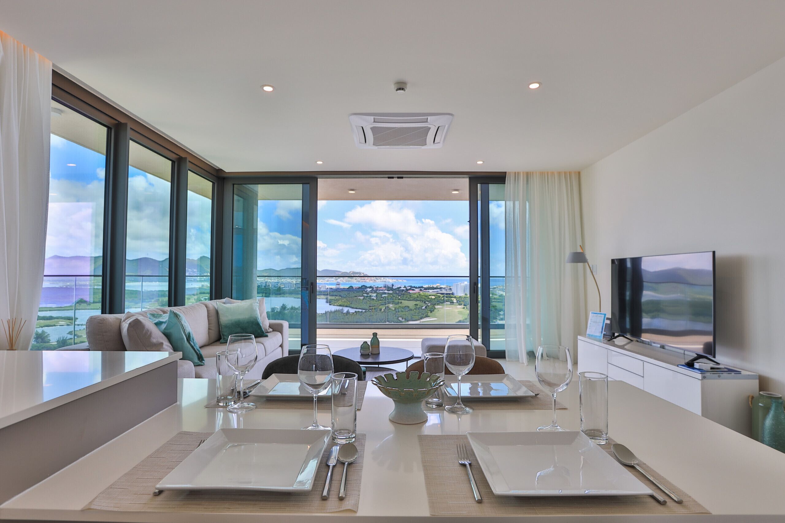 A modern dining area with a table set for two, large windows with a view of the landscape, and a television on the right.