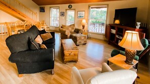 Een ruime woonkamer met houten vloeren, een zwarte bank, een witte fauteuil en een houten salontafel. Er is een trap met een houten leuning die naar een hoger gelegen niveau leidt.