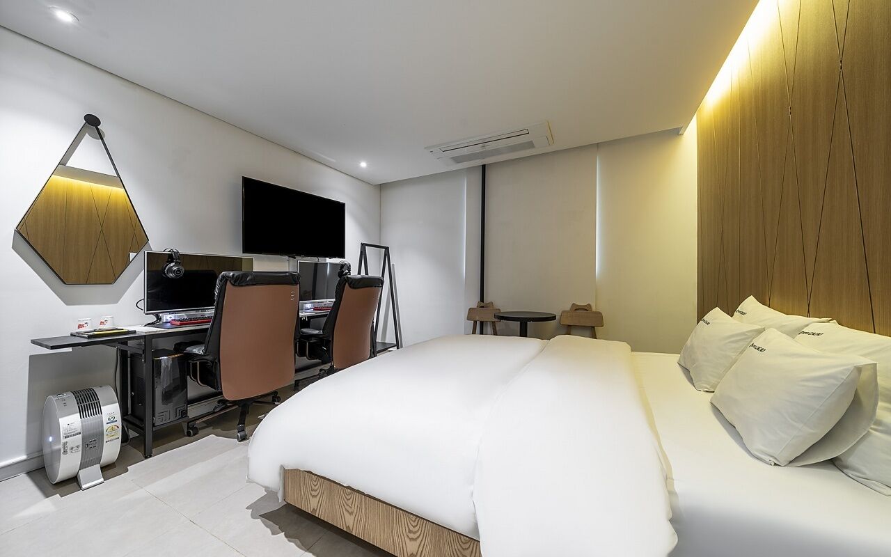 Una habitación de hotel moderna con una cama grande, un escritorio con computadora, una silla, un televisor y una lámpara de techo hexagonal única.
