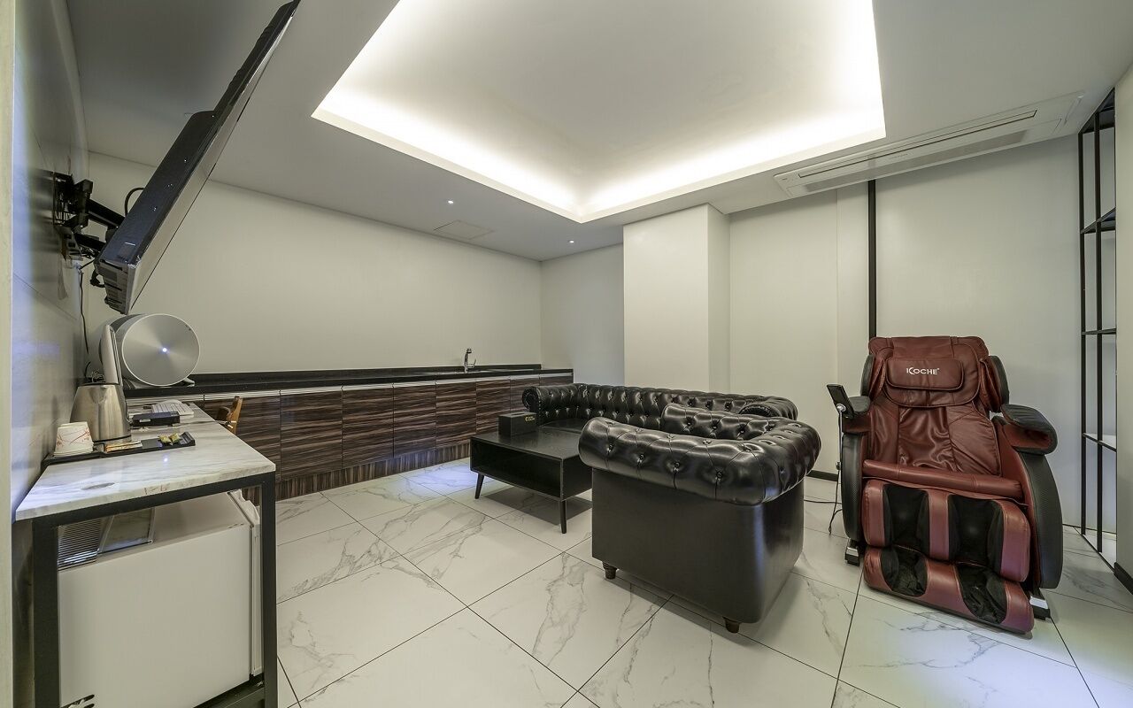 Una habitación moderna con sillón de masaje, sofá, escritorio con lámpara y ventana grande.