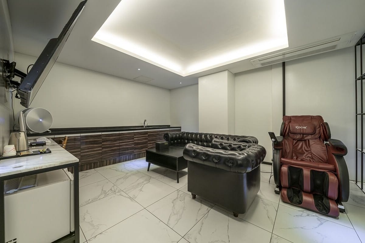 Una habitación moderna con sillón de masaje, sofá, escritorio con lámpara y ventana grande.