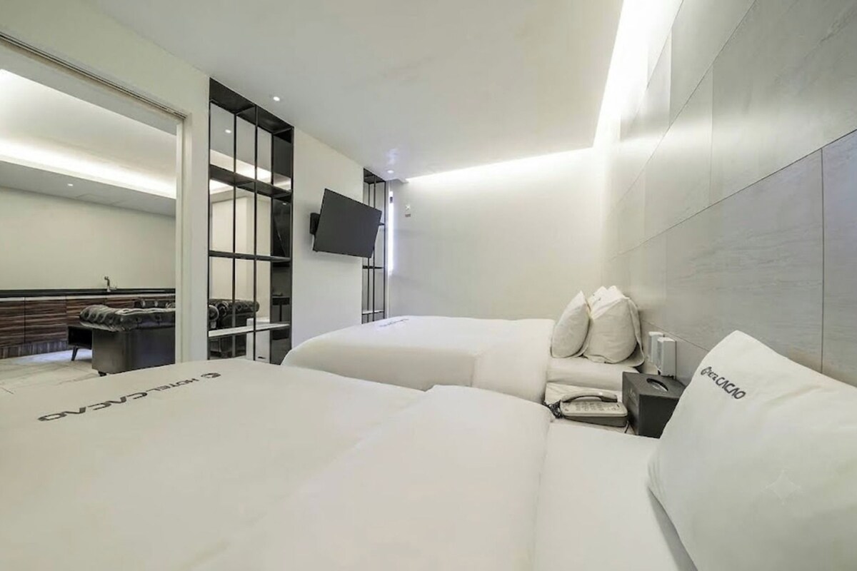 A modern hotel room with two beds, a flat-screen TV, and a bathroom visible through a glass door.