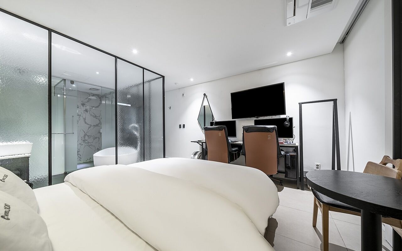 Habitación de hotel moderna con ducha de vidrio, cama con ropa blanca, escritorio con computadora y televisor de pantalla plana montado en la pared.