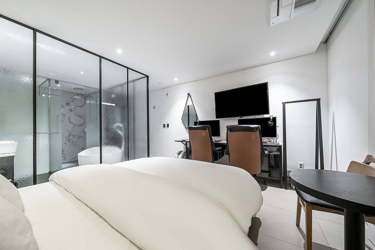 Moderni hotellihuone, jossa on lasiseinällä varustettu suihku, valkoisilla tekstiileillä varustettu sänky, työpöytä tietokoneella ja seinälle asennettu litteänäyttöinen televisio.