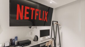 Een moderne kamer met een grote flatscreen-tv aan de muur, waarop in rode letters 'NETFLIX' staat. Daaronder een bureau met een computer, toetsenbord, muis en koffiezetapparaat.
