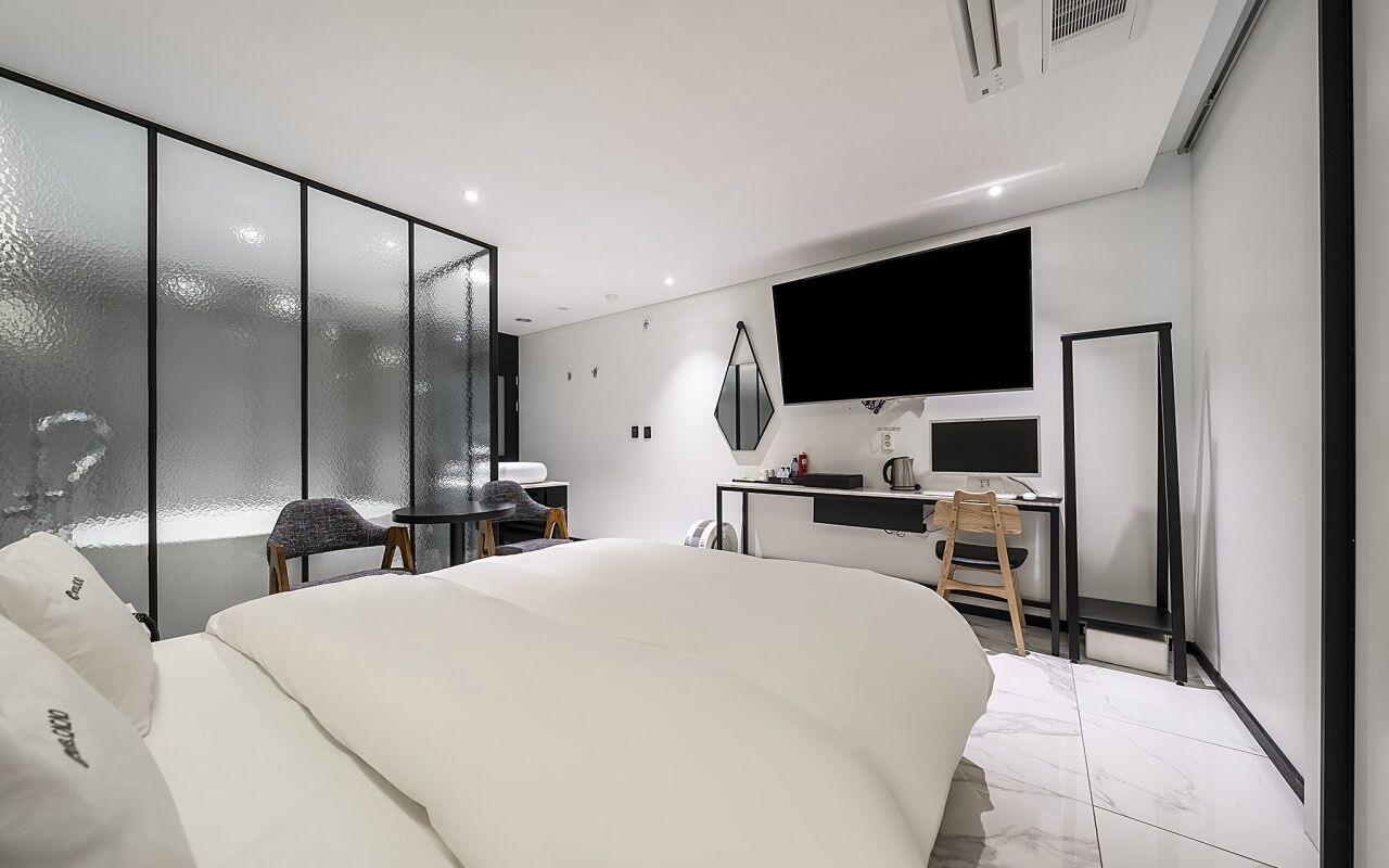 Una habitación de hotel moderna con una cama grande, un escritorio con una computadora, un televisor y una pared divisoria de vidrio.