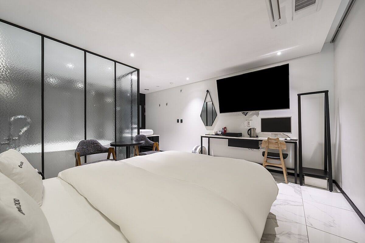 Moderni hotellihuone, jossa on suuri sänky, työpöytä tietokoneella, televisio ja lasiseinä.