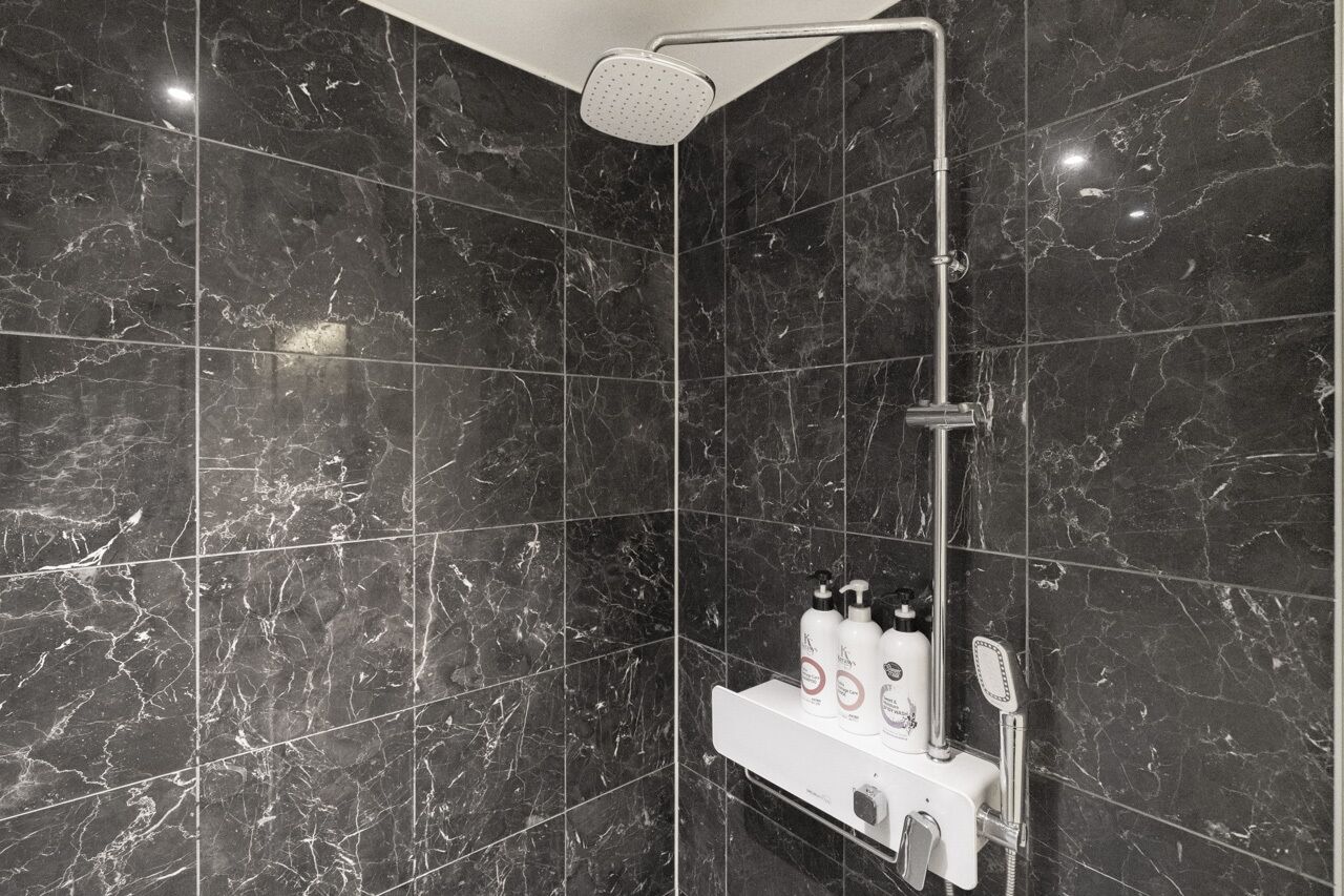 A modern bathroom with dark marble tiles, a showerhead, and a shelf with toiletries.