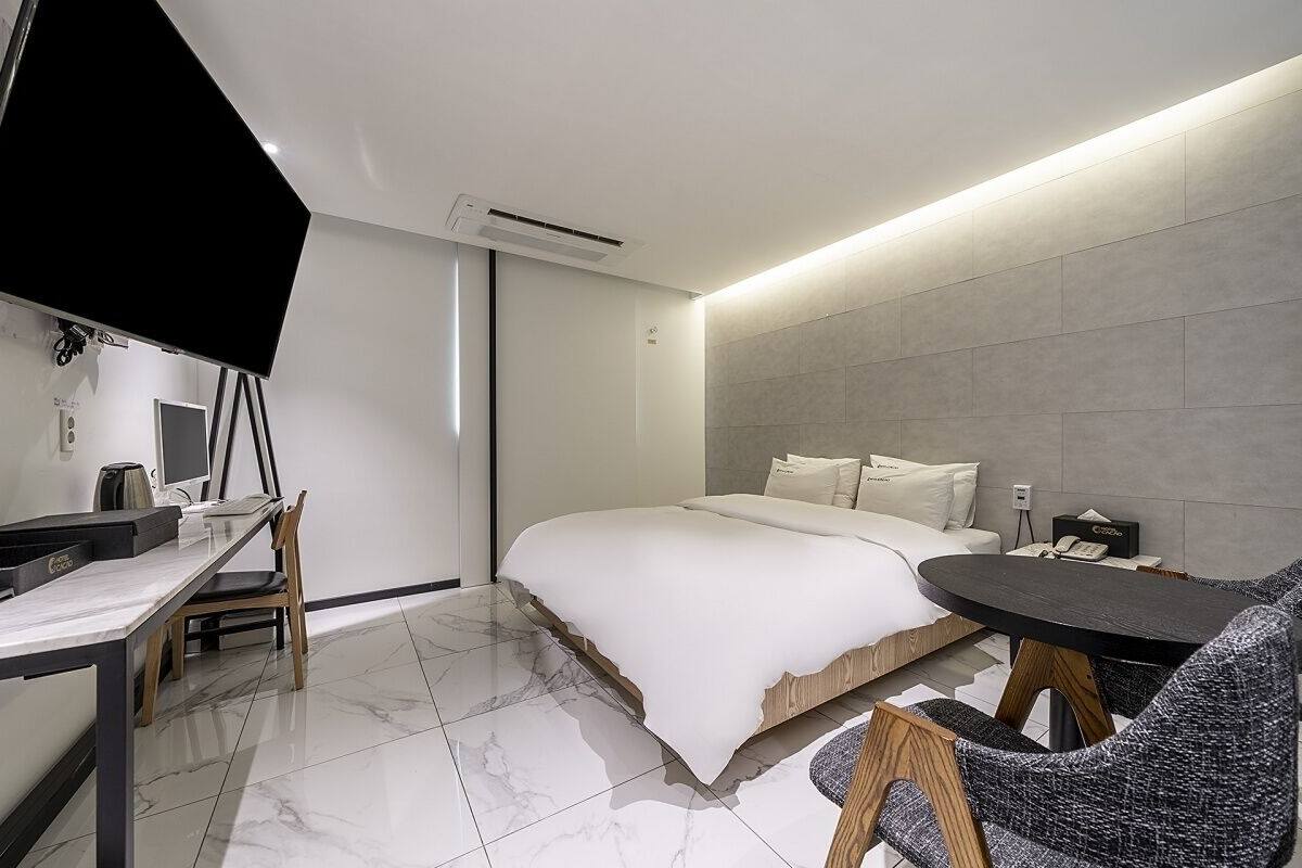 Moderni hotellihuone, jossa on sänky, työpöytä tietokoneella, tuoli ja pieni pöytä.