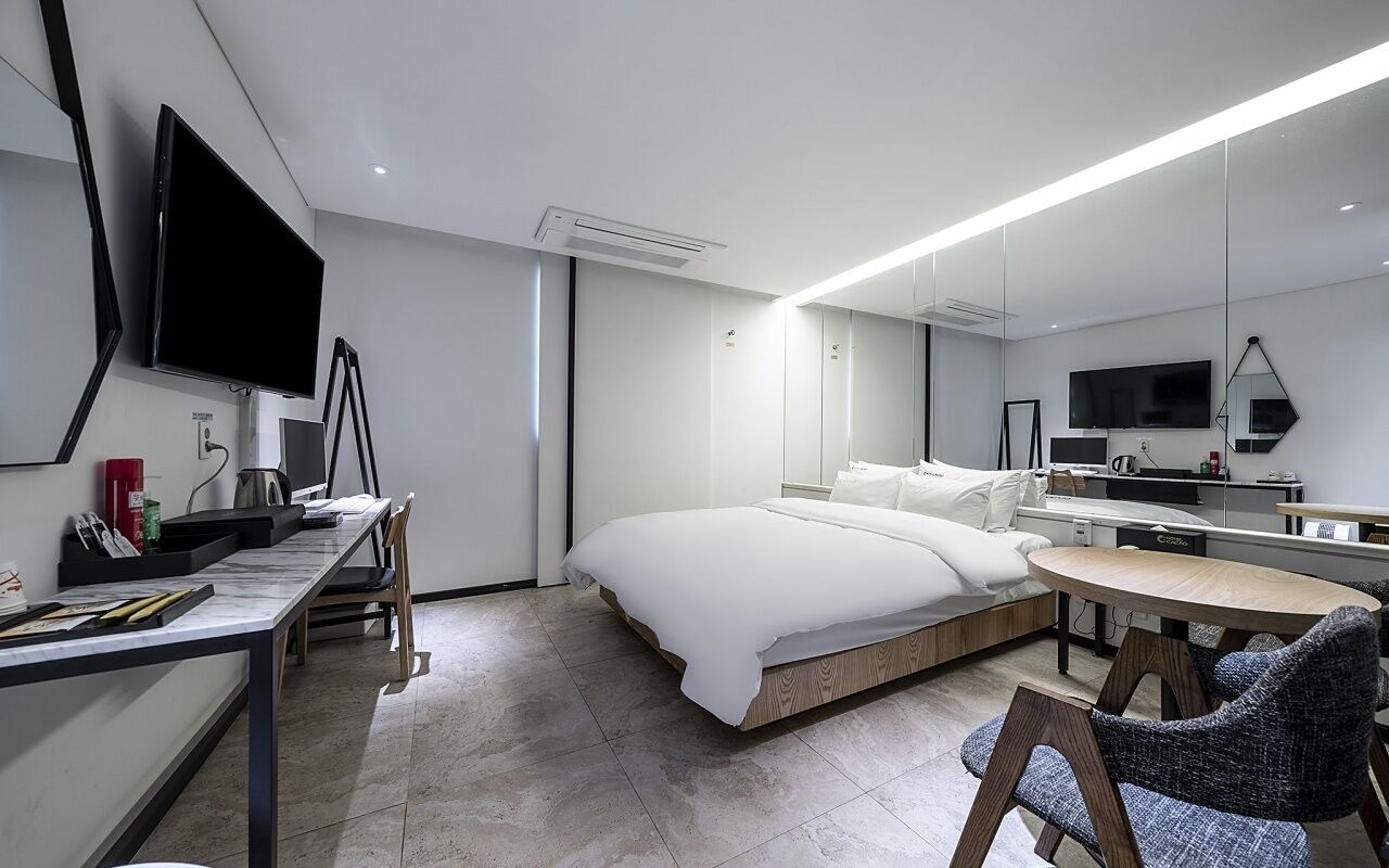 Una habitación de hotel moderna con cama, escritorio, silla, televisor y un espejo grande.