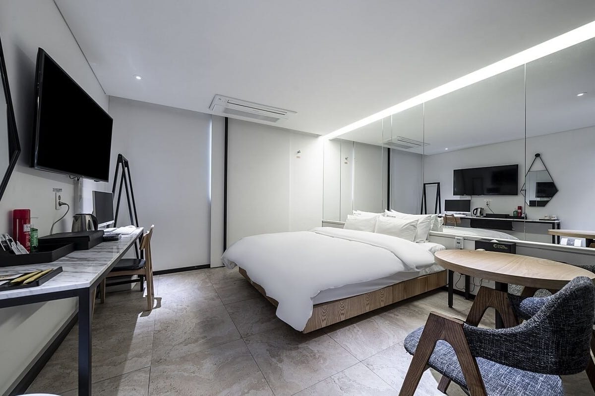 Una habitación de hotel moderna con cama, escritorio, silla, televisor y un espejo grande.