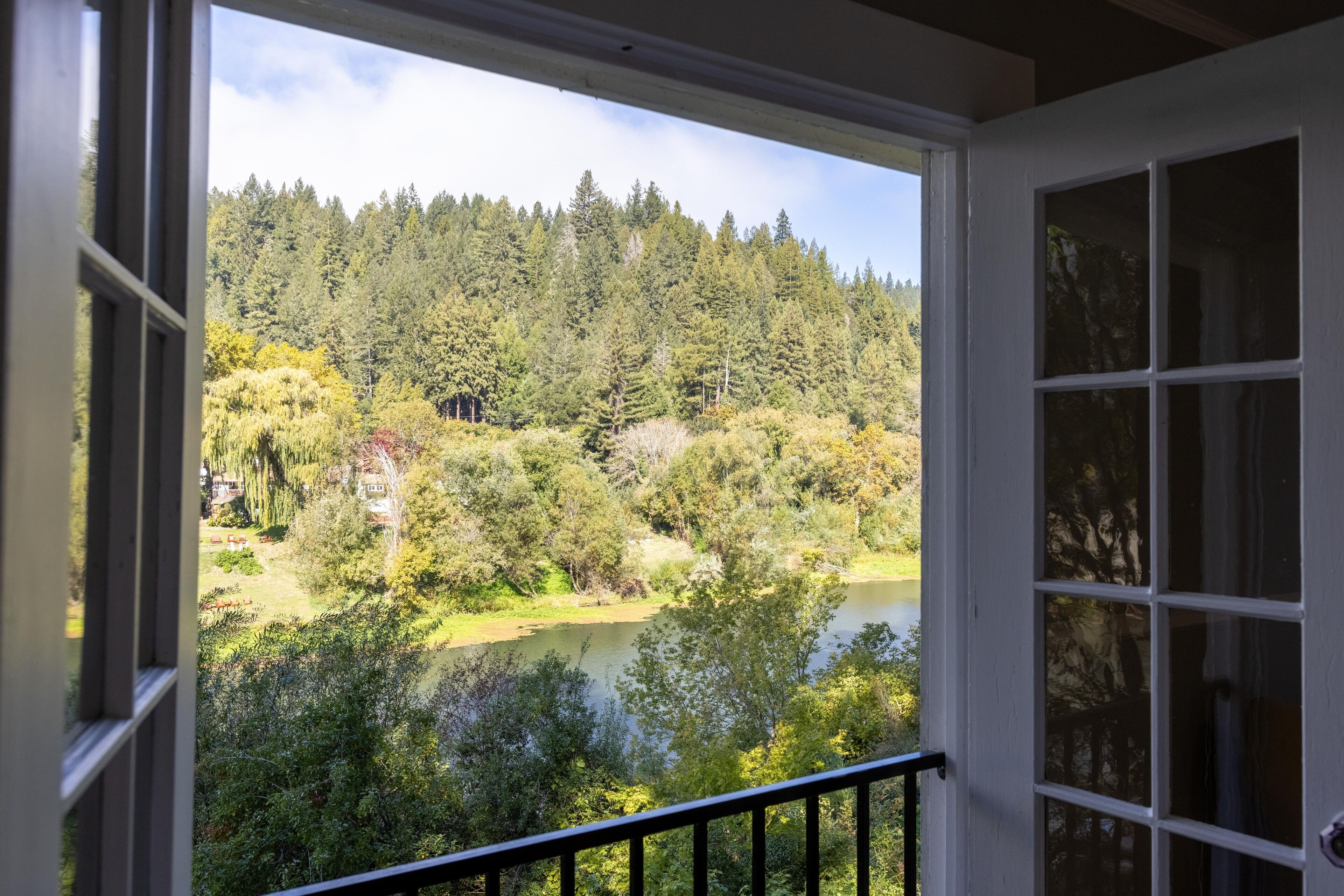 Une vue depuis une fenêtre dotée d’un cadre blanc et d’une balustrade noire, donnant sur une zone boisée avec un lac et une colline en arrière-plan.