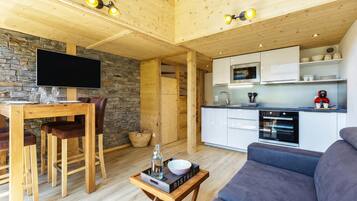 Un intérieur moderne avec une table à manger en bois, des chaises, un canapé, un coin cuisine doté de meubles blancs et un mur en pierre.