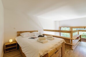 A bedroom with a wooden bed, white bedding, a nightstand with a lamp, and a view of greenery through a window. - Grand Roc A217 150m from Slopes, Mont-Blanc View, Parking & WiFi, Argentière (Chamonix), France (Argentière (Chamonix))