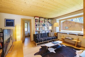 A cozy living room with a wooden ceiling, a bookshelf, a sofa, a TV, and a window with a view of a wooden cabin.