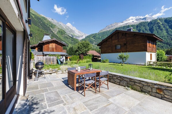 A patio with stone flooring, a wooden table, and chairs, overlooking a mountainous landscape with wooden buildings.