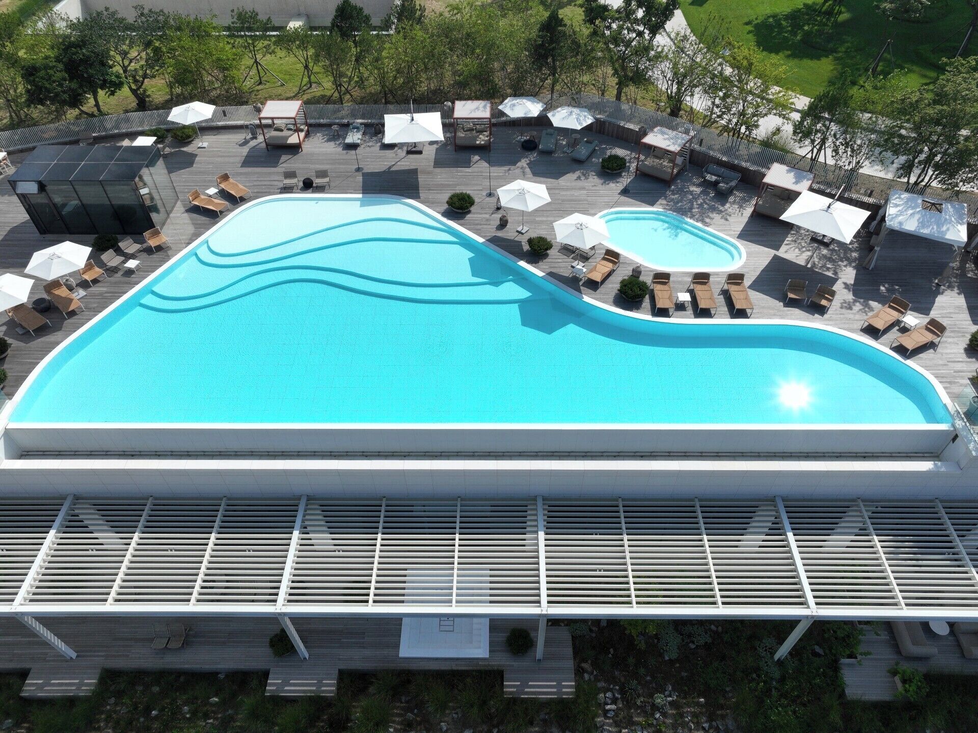 A large swimming pool with sun loungers and umbrellas, surrounded by a wooden deck and a covered seating area.