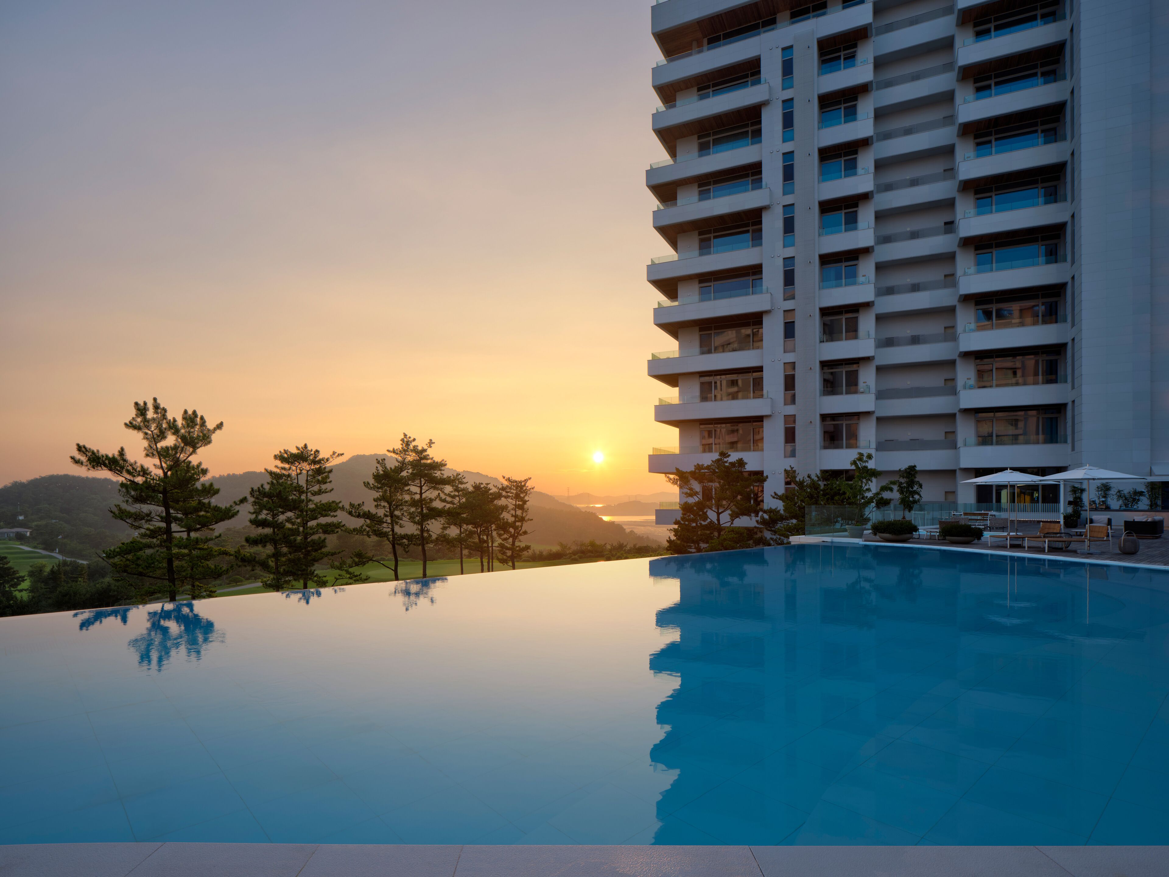 Un hotel moderno con piscina que refleja el atardecer, rodeado de árboles y vistas a colinas distantes.