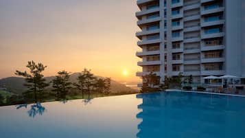 Un hotel moderno con piscina que refleja el atardecer, rodeado de árboles y vistas a colinas distantes.